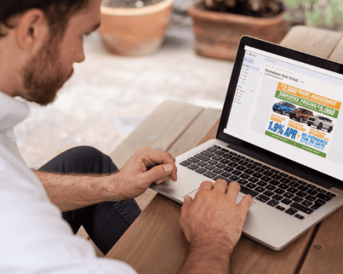 Man at wooden table using laptop displaying car dealership ad featuring vehicle images and promotional offers, including trade-in assistance and low APR financingβtargeted marketing for Willowood Ventures automotive sales.