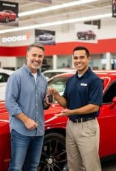 Customer receives car keys from dealership employee in front of a red Dodge vehicle inside showroom, showcasing Willowood Ventures’ automotive services. Dodge branding and MichelleWP logo visible in background.