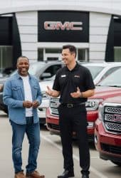 Two men, one in a black GMC shirt and the other in a denim jacket, smile and chat in front of multiple parked GMC vehicles at a Willowood Ventures dealership. The dealership building and prominent GMC signage are visible behind them.