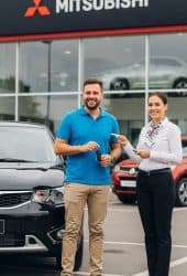 Saleswoman handing car keys to customer outside Mitsubishi dealership, showcasing new vehicle handover with dealership building and cars in background—ideal for Willowood Ventures automotive marketing.
