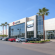 Modern car dealership showroom with glass facade and palm trees, showcasing multiple new vehicles; digital display above entrance features a car in motion, under clear blue sky—ideal for Willowood Ventures automotive marketing.