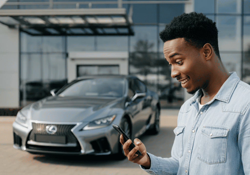 Young man in denim shirt stands outside a modern building, smiling at his smartphone with a grey Lexus parked behind him, highlighting Willowood Ventures’ latest automotive BDC promotions.