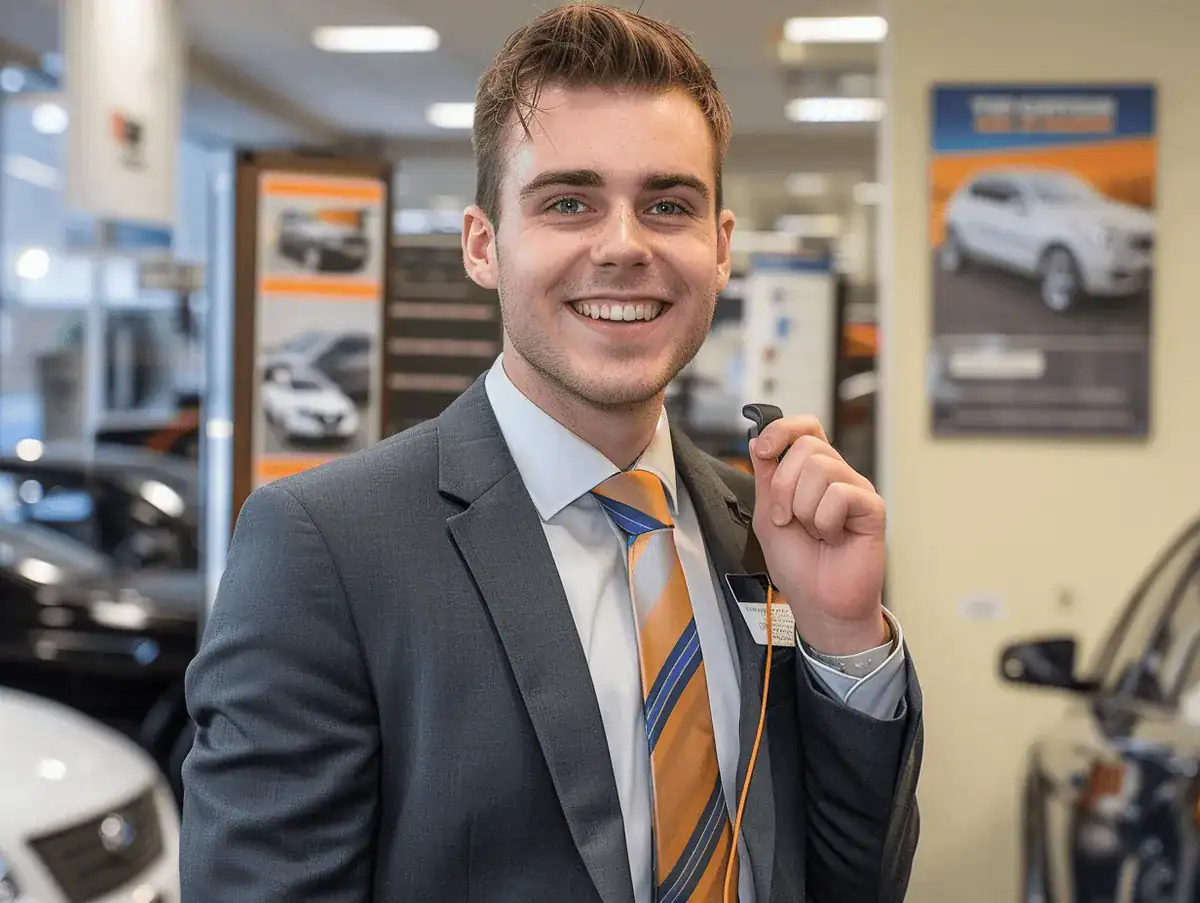 Smiling car dealership salesperson holding keys in a showroom.