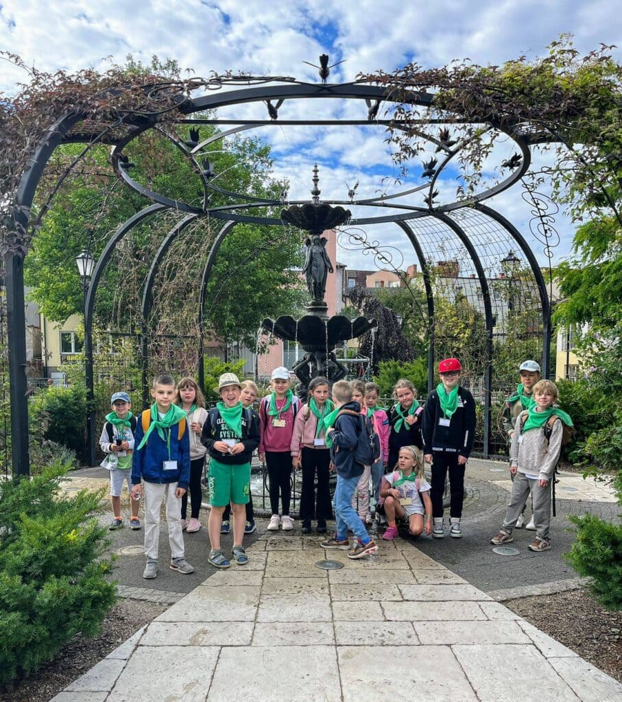 Dzieci na wycieczce szkolnej w ogrodzie z fontanną i metalową konstrukcją, otoczone zielenią i architekturą, uczestniczą w aktywności edukacyjnej na świeżym powietrzu, promując zabawę i naukę.