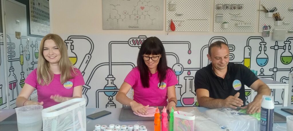 Kolorowa, naukowa atmosfera w laboratorium, z trzema osobami rozmawiającymi i pracującymi przy eksperymentach, chemiczne probówki i narzędzia na stole, edukacyjne grafiki na ścianie, logo Wittlab.