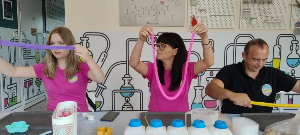 Kolorowe eliksiry chemiczne eksperymenty naukowe w laboratorium, uczestnicy w pinkowych koszulkach, prezentacja próbek i ciekawych substancji, nauka chemii dla dzieci i dorosłych, edukacja i zabawa naukowa.