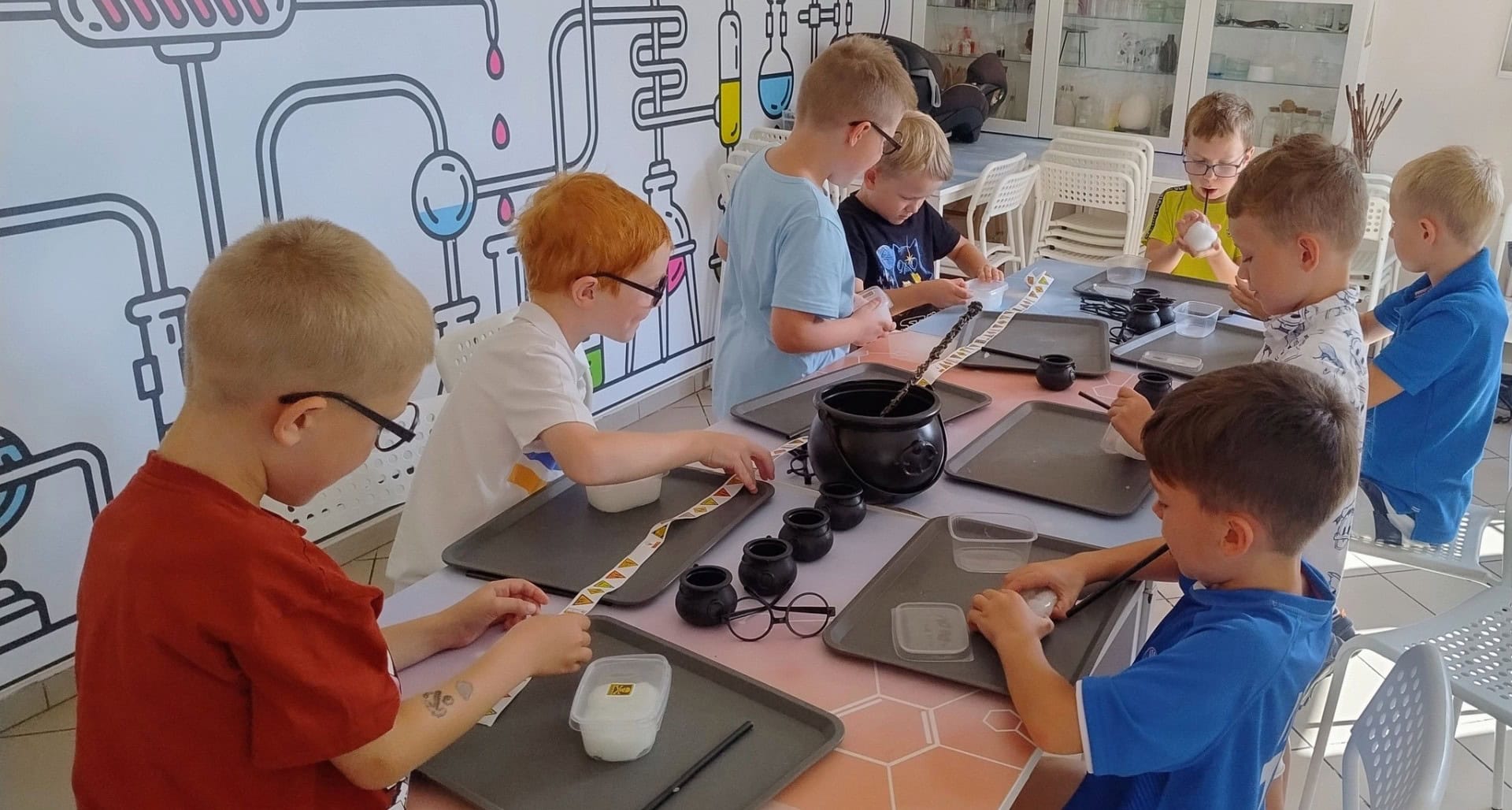 Kolorowa sala warsztatowa z dziećmi uczestniczącymi w kreatywnych zajęciach naukowych, wyposażona w naczynia i materiały do eksperymentów chemicznych, ze ścianami ozdobionymi motywami laboratoryjnymi.