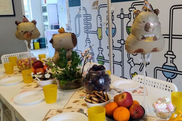 Kolorowa dekoracja urodzinowa z balonami w kształcie słodkich zwierząt, owocami i słodyczami na stole, idealna na przyjęcie dla dzieci, z nowoczesnym motywem naukowym na ścianie.
