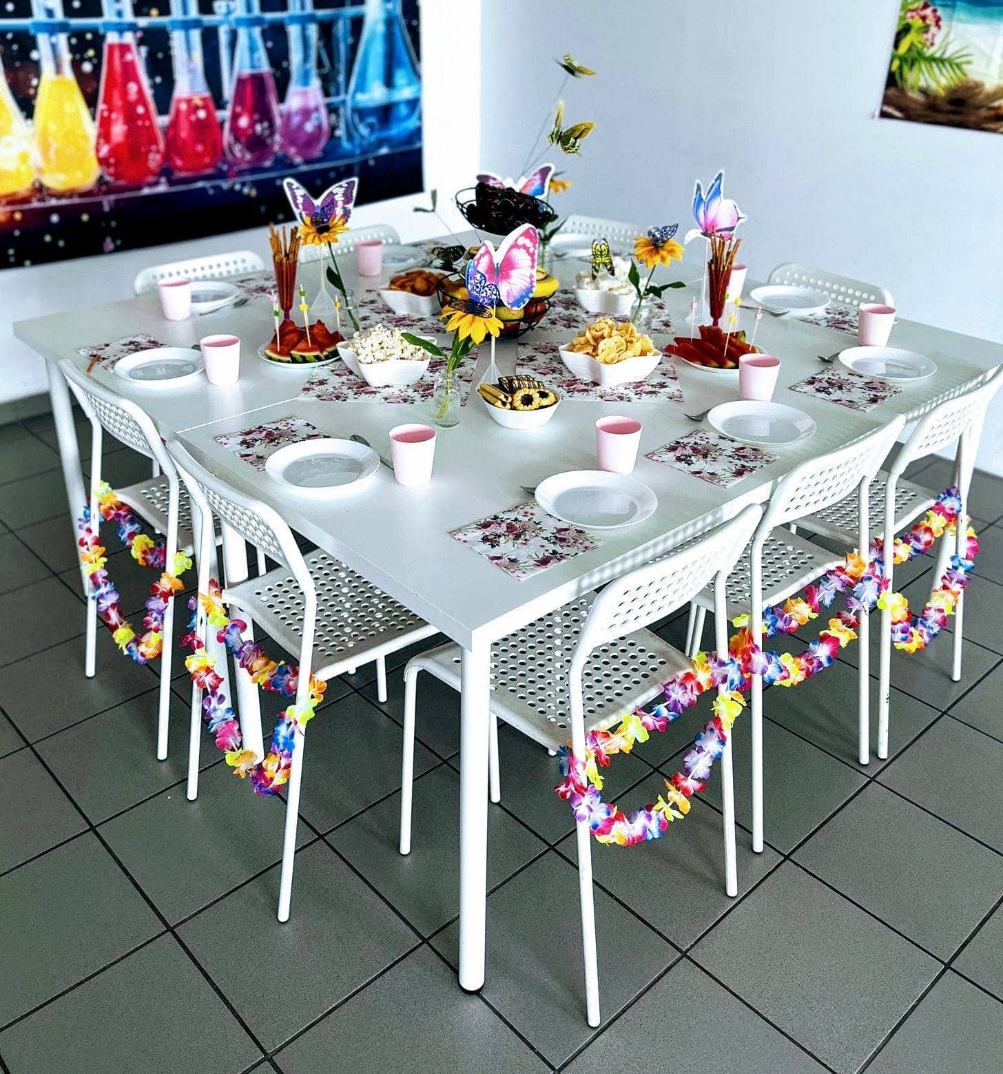 Kolorowa dekoracja na przyjęcie dla dzieci, kwiatowe i motylkowe motif, na białym stole z talerzami, kubkami i transparentami, idealna na dziecięce urodziny lub imprezy tematyczne.