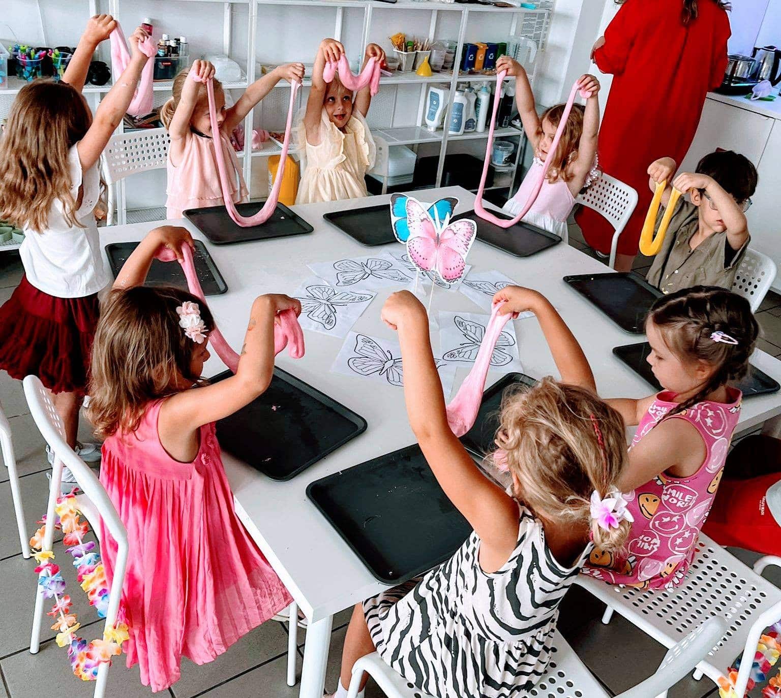 Kolorowe dzieci podczas warsztatów plastycznych z rozciąganiem masy plastycznej, ubrane w modne stroje, wokół stołu z papierami do malowania i maskotkami motyli, kreatywna zabawa w przyjaznym środowisku.