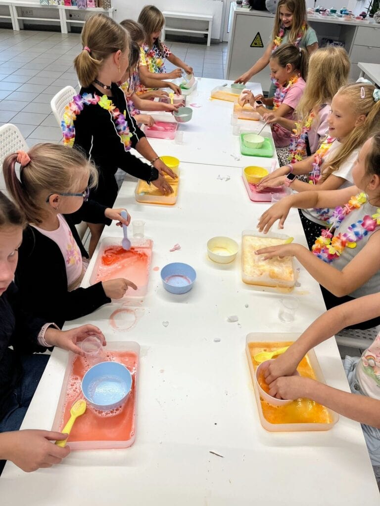 Kolorowa grupa dzieci podczas warsztatów z tworzenia slime'u, w kuchni szkolnej, z uśmiechami i zaangażowaniem w lepienie masy plastycznej.