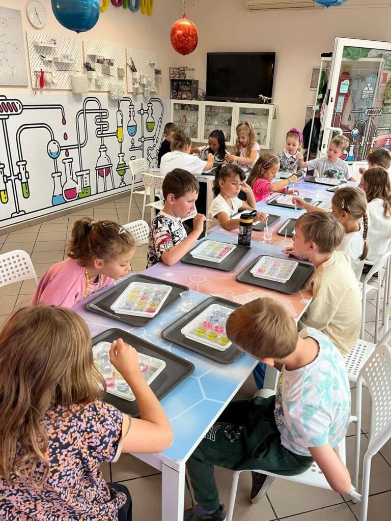 Kolorowa sala do warsztatów dla dzieci z chemicznymi eksperymentami, pełna uśmiechniętych uczestników, z ciekawym grafiką naukową na ścianie, kreatywne zajęcia edukacyjne, idealne dla rozwoju małych naukowców.