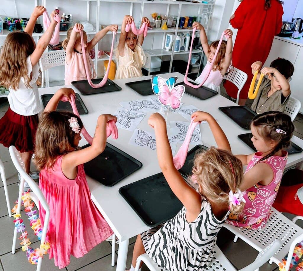 Kolorowe, kreatywne zajęcia plastyczne dla dzieci w wiej szkole, dziewczynki i chłopcy trzymają różowe i żółte rozciągnięte plasteliny podczas warsztatów artystycznych, stół z rysunkami motyli w tle.