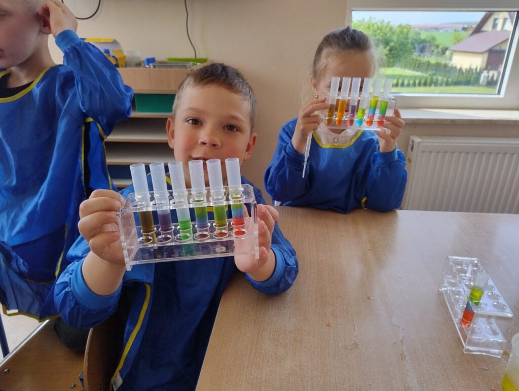 Kolorowe testy naukowe przeprowadzane przez dzieci w edukacyjnym laboratorium, pokazujące fascynację chemią i nauką, w naturalnym świetle przy oknie, z uśmiechami i ciekawością.