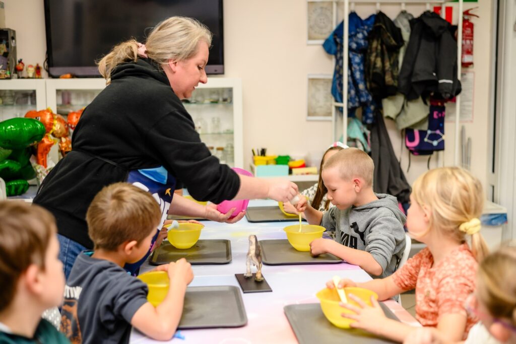 Chłopiec i dziewczynki biorą udział w warsztatach kulinarnych dla dzieci, uczą się gotować pod nadzorem instruktorki, w przyjaznym i kolorowym otoczeniu kuchni lub szkoły.