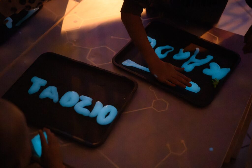 Tablica świetlna z napisem "TAOZIC" tworzone na tablecie, uczestnicy korzystają z interaktywnych narzędzi w ciemnym pomieszczeniu, promując kreatywność i innowacyjne rozwiązania w digital art.