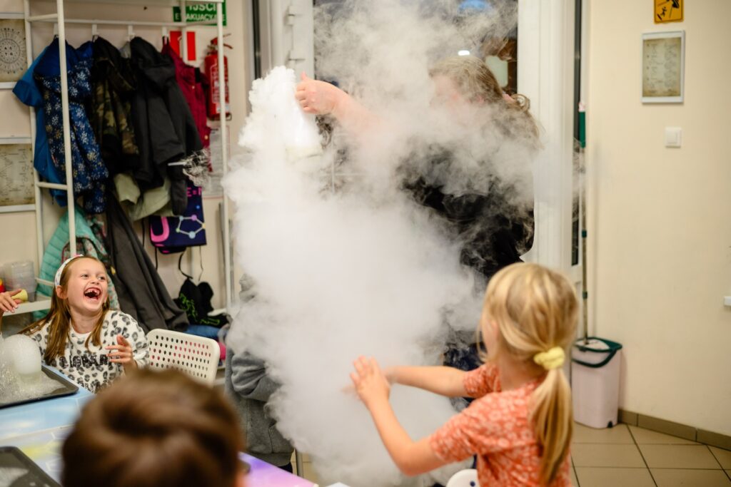 Radosne dzieci uczestniczące w eksperymencie naukowym, obserwujące dym słoneczny w laboratorium Wittlab, inspirujące do nauki i odkrywania tajemnic fizyki i chemii.