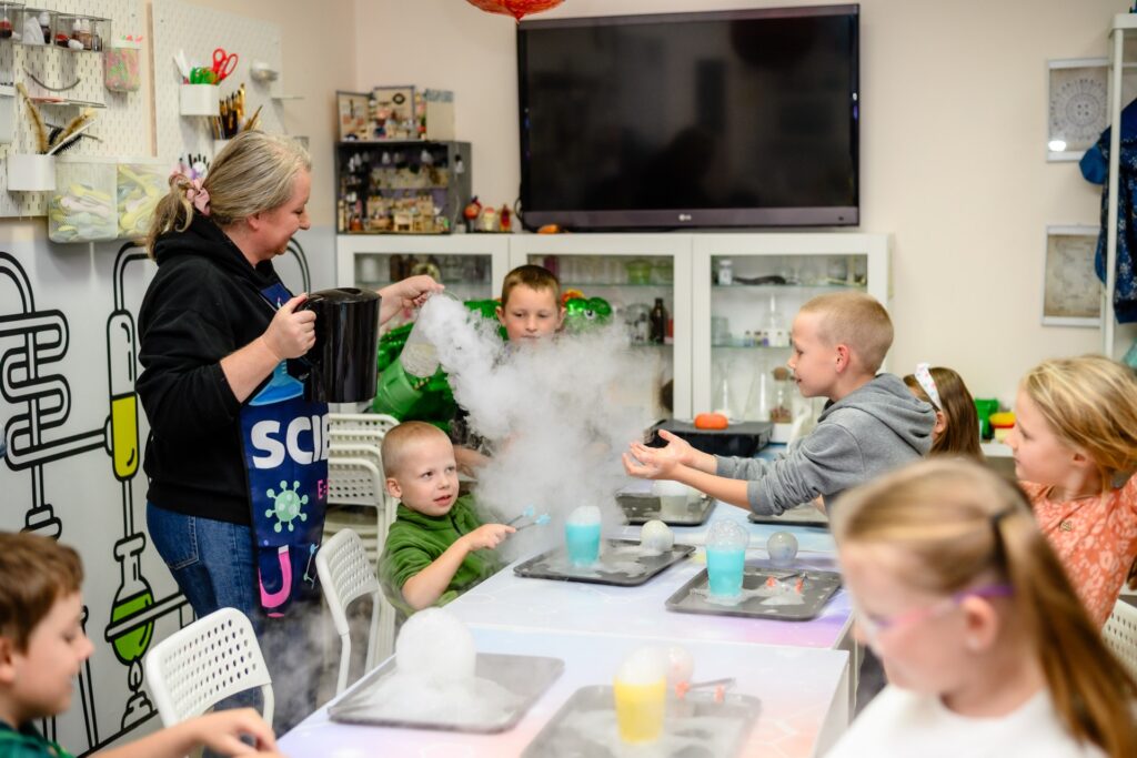 Kolorowa animacja chemiczna z dziećmi i instruktorem podczas eksperymentu z znikającymi dymami na tle edukacyjnej pracowni naukowej.