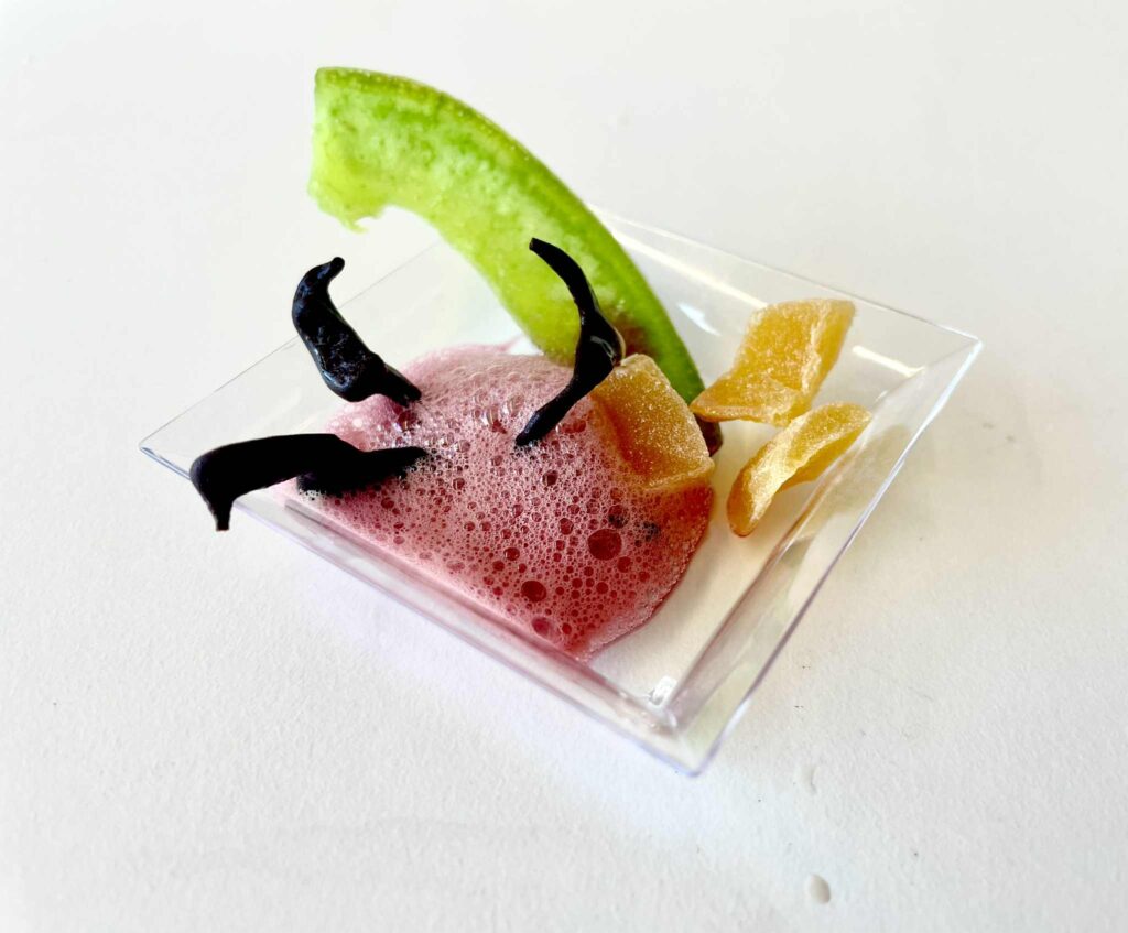 Alt text: Koktajl z różowym musem, plasterkiem limonki, kawałkami kandyzowanego imbiru i czarnymi przyprawami w małym kieliszku, na białym tle.