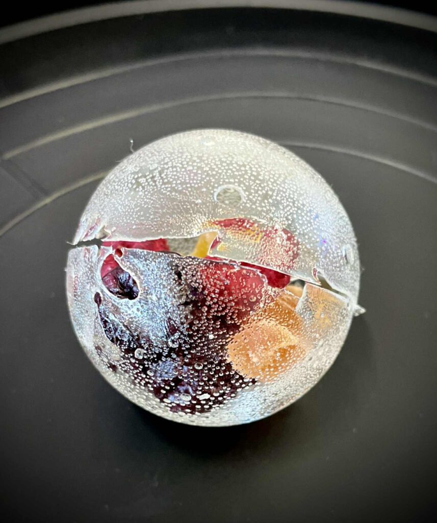 Kula lodowa z owocami i napojem gazowanym w środku, zamknięta w kryształowym lodzie, prezentacja deseru lub drinka, elegancki i efektowny sposób podania napoju, idealna na specjalne okazje, foto z bliska, widok z góry.