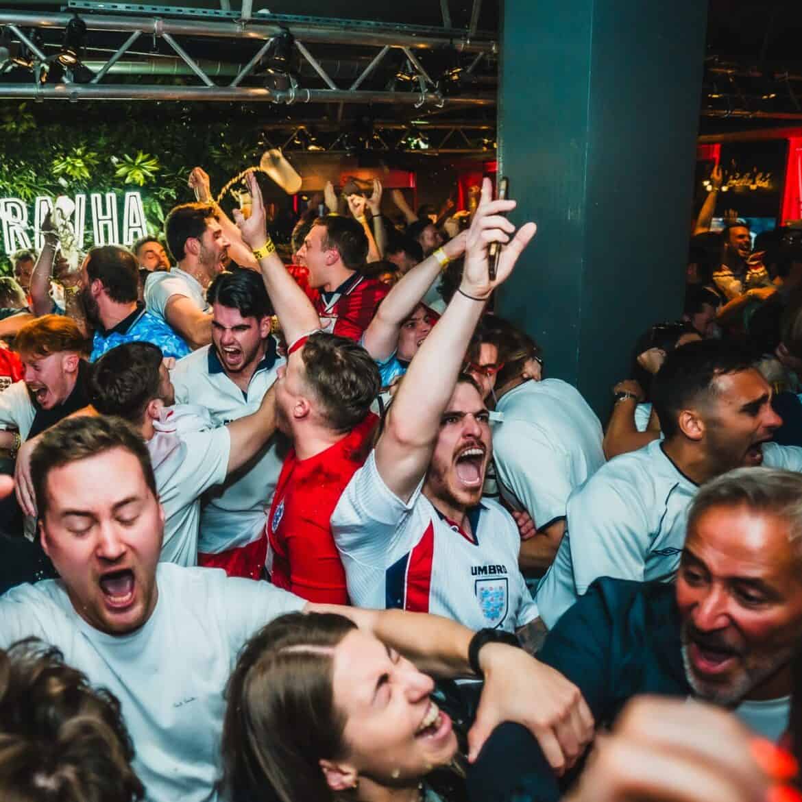 Packed crowd of England football fans erupting in celebration at Belushi's Shepherd's Bush after a World Cup goal, with confetti in the air and the Pravha bar area in the background