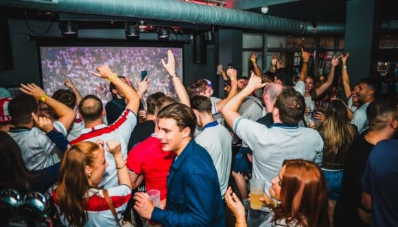 England fans celebrating a goal during a World Cup 2026 watch party at Belushi's sports bar in London, with crowds cheering and hands raised in front of a large indoor screen