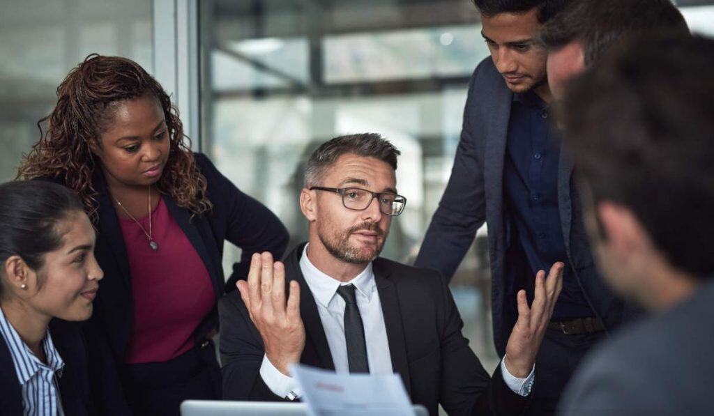 Confident businessman explaining ideas during team meeting, diverse professionals collaborating on project strategy at modern office.