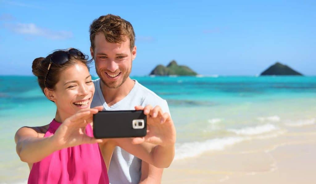 Vibrant couple taking a selfie on a sunny beach in Australia, with turquoise water and tropical islands in the background, showcasing travel and holiday experiences.