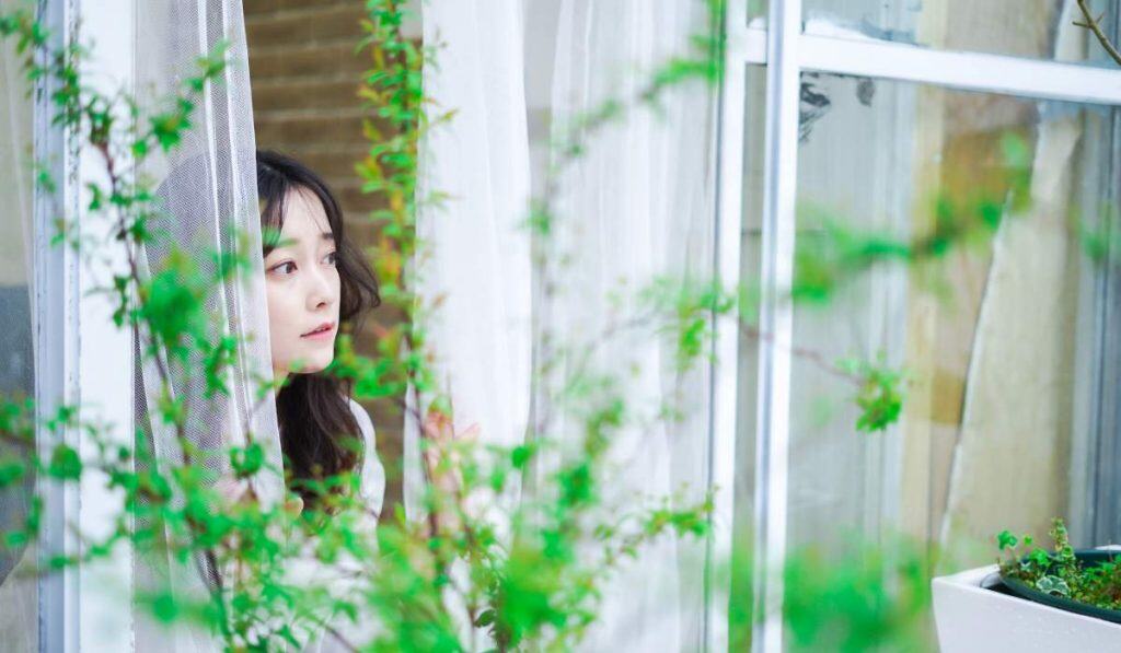 Lush green plants and a young woman looking out from a bright, airy conservatory with white curtains and large windows at World Lee Journeys, promoting travel, relaxation, and lifestyle experiences.