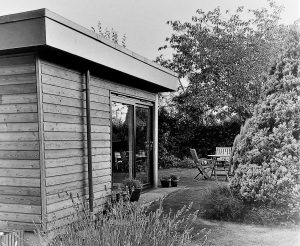 Bright wooden garden room with glass door and outdoor seating area, surrounded by lush greenery.