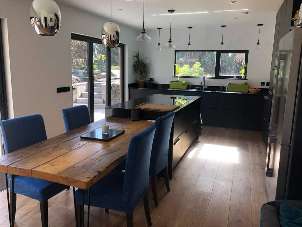 Contemporary kitchen with black cabinets, wooden dining table, and large windows for natural light.