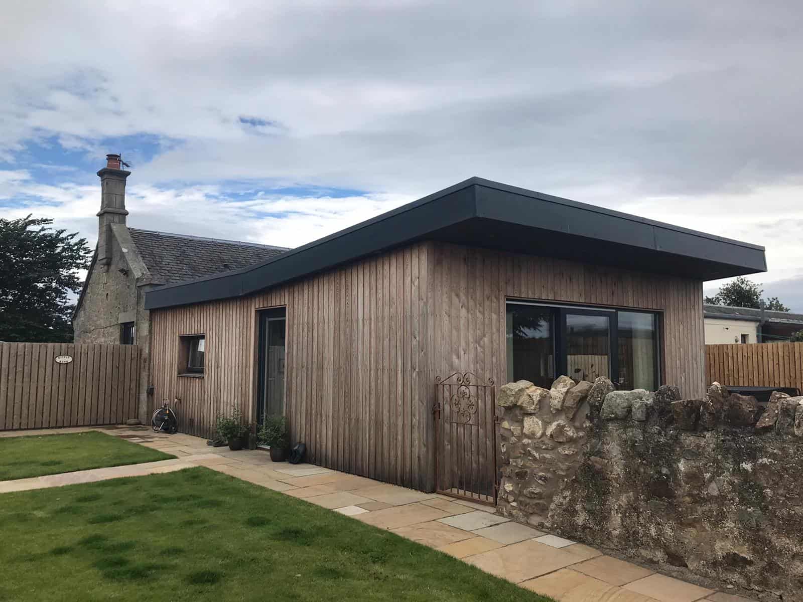 Alt: Contemporary wooden house extension with large glass doors and stone wall, in a garden setting.
