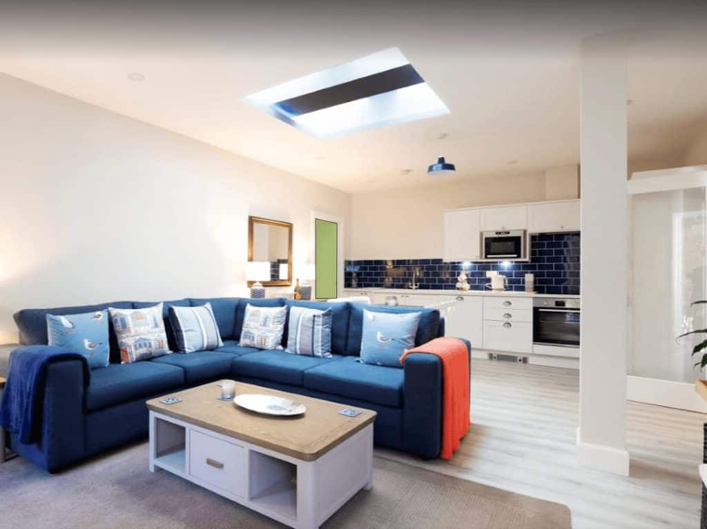 Bright living area with large navy sofa, colourful cushions, and seamless kitchen with blue tiles and built-in appliances.