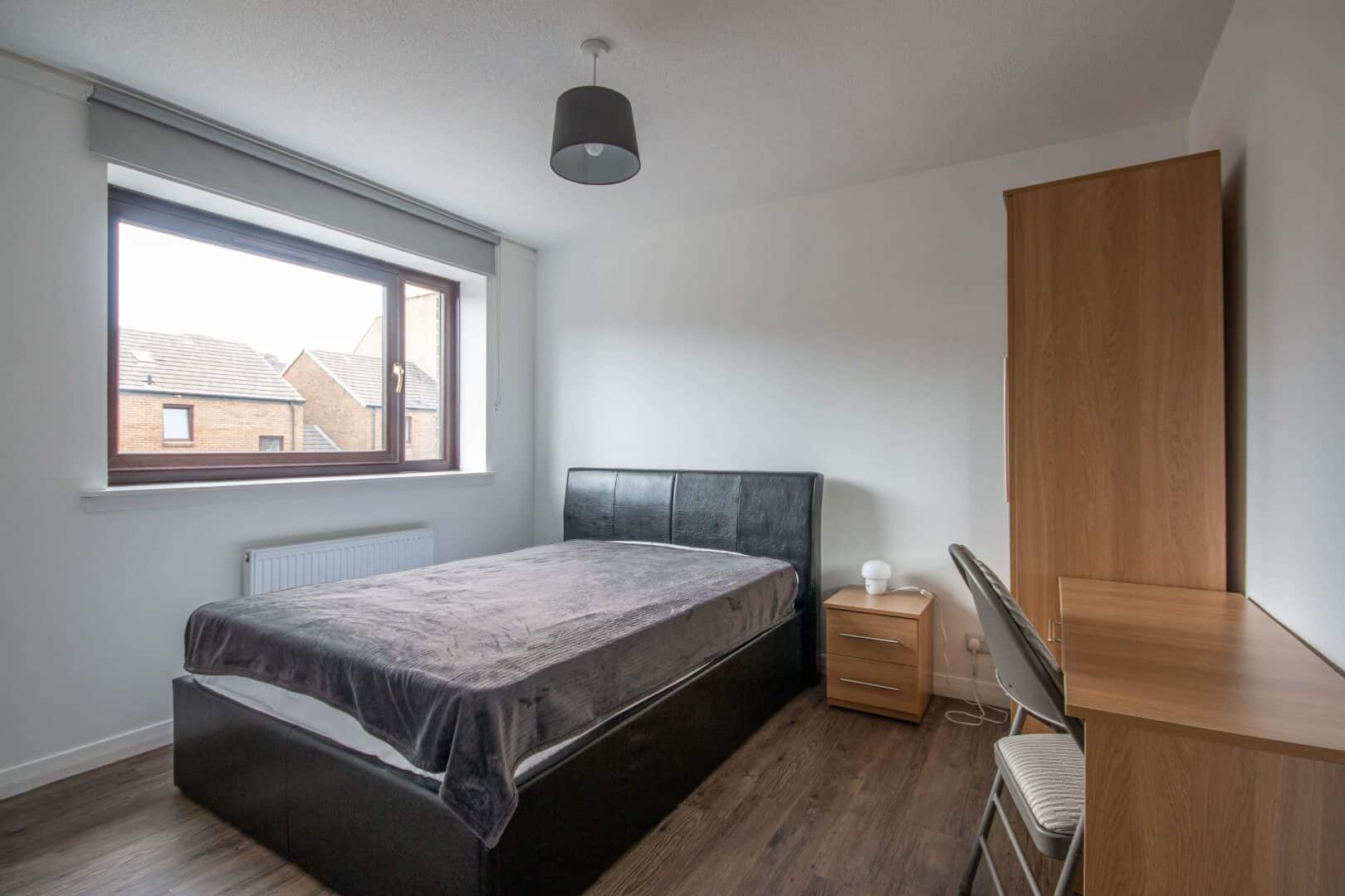 Comfortable guest bedroom featuring a wooden wardrobe, desk, and large window showcasing neighbourhood rooftops.