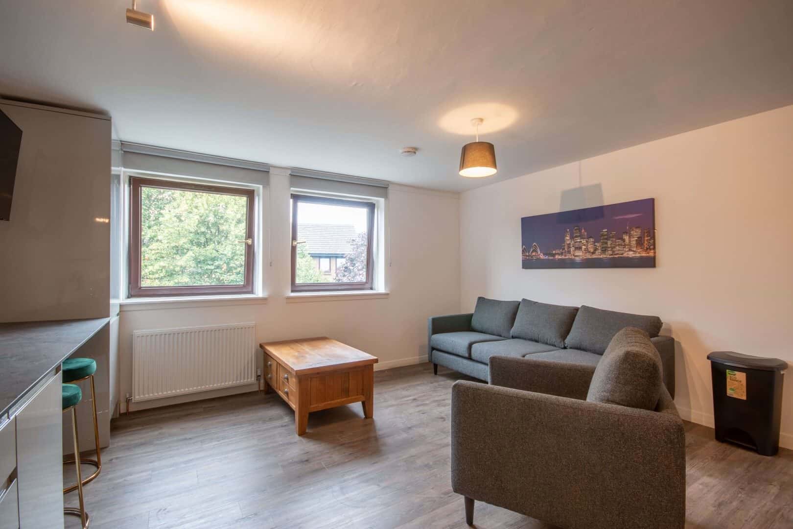 Bright living room with dual large windows, grey sofa, wooden coffee table, and city skyline wall art, reflecting stylish residential architecture.