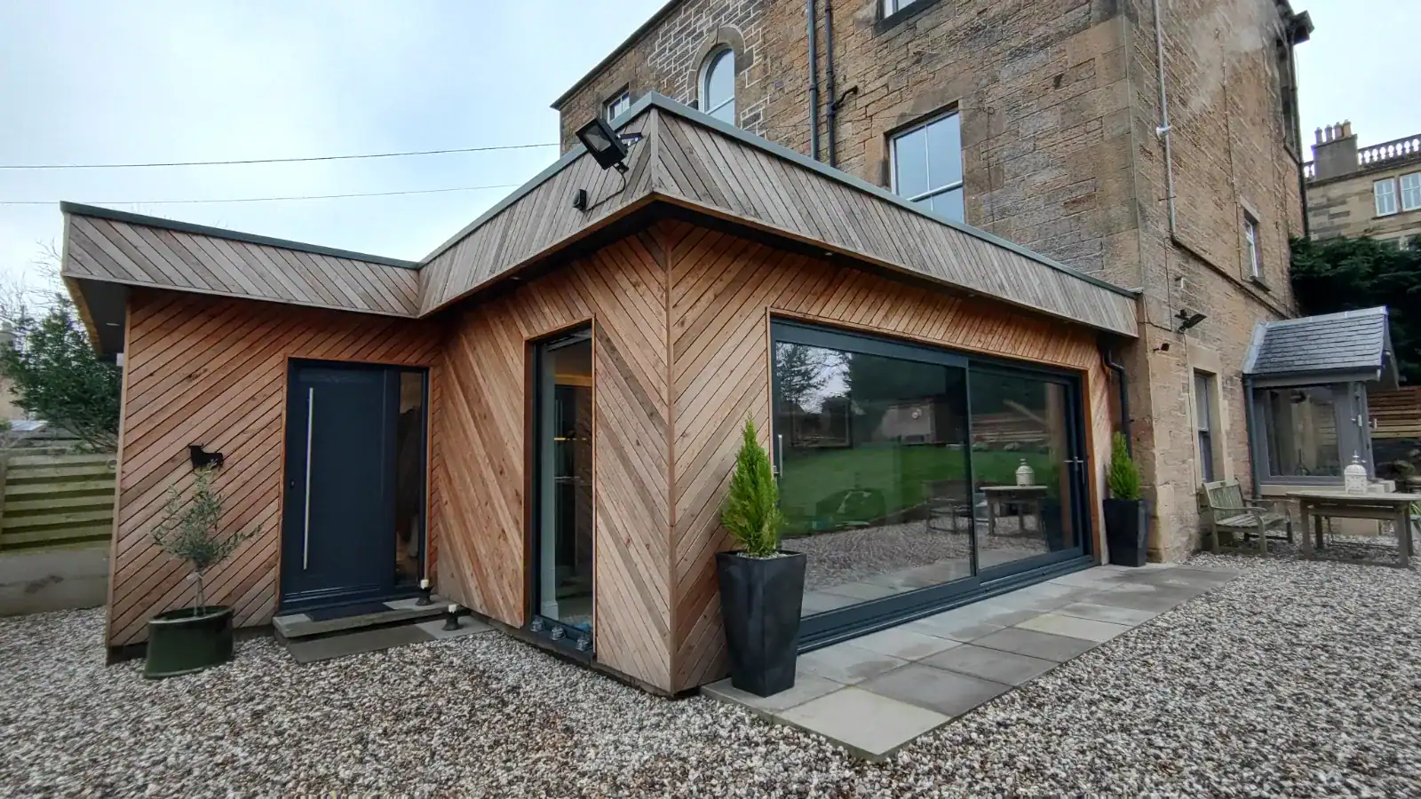 Modern wooden home extension designed by ABC Architecture with large glass doors, outdoor seating, and potted plants.