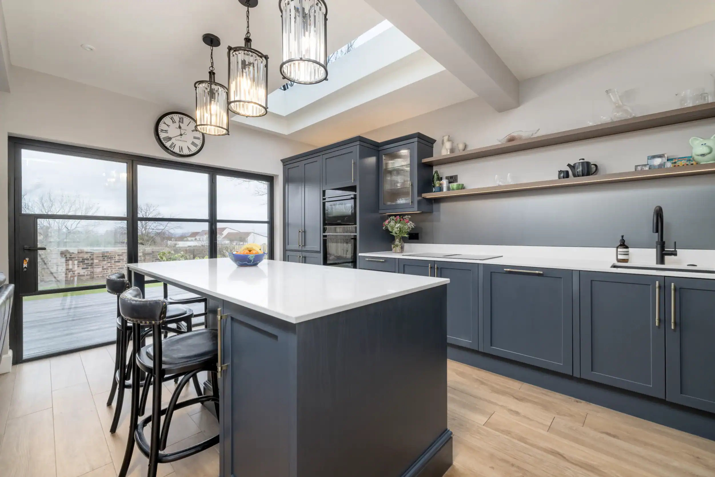 Bright and stylish kitchen featuring blue cabinetry, white countertops, large windows, and open shelving for functional elegance.