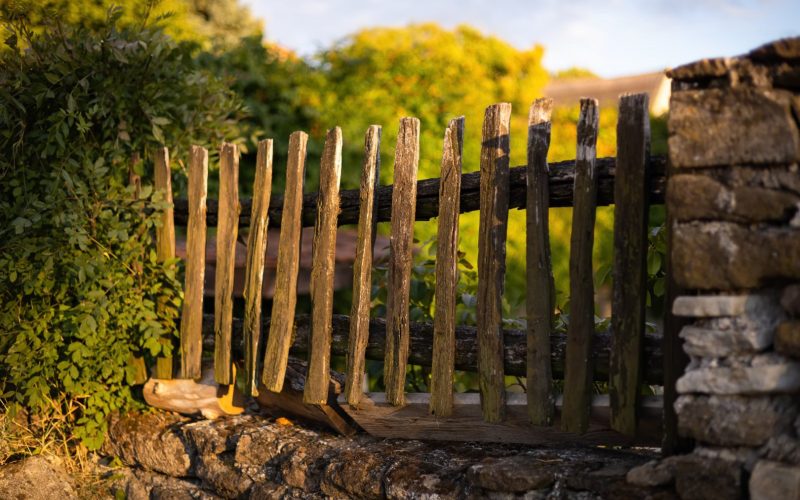 Old wooden picket fence built into a wall from stones. Sunset over an aged timber barrier protecting privacy of home on a village. Hardwood brown material.