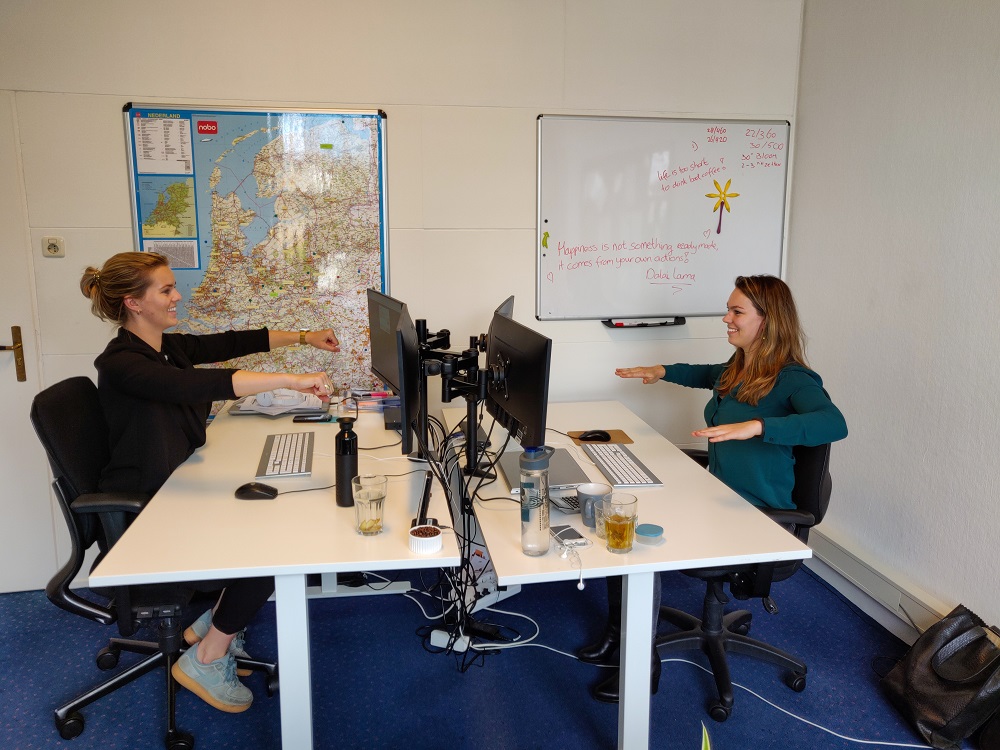 Medewerkers doen de oefening double-arm row achter hun bureau. Ze zitten en steken de armen naar voren, waarna ze deze terugtrekken naast hun lijf met de armen 90 graden gebogen.
