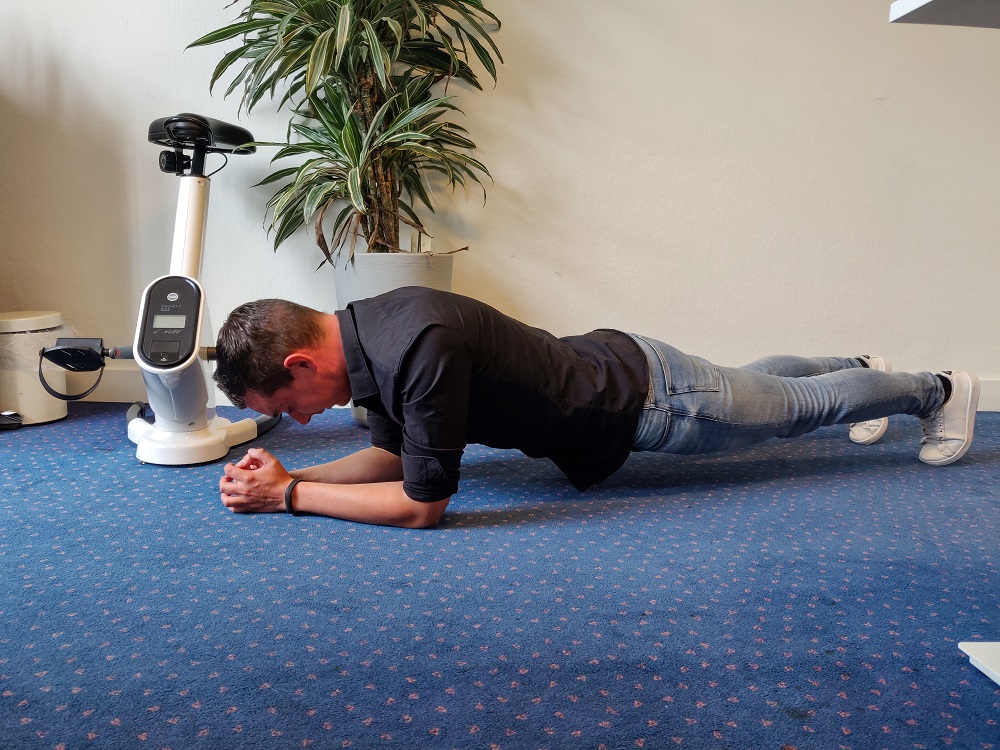 Medewerker doet de oefening planking. Hij leunt op onderarmen en tenen en heeft de rest van het lijf van de grond getild. Recht als een plank.