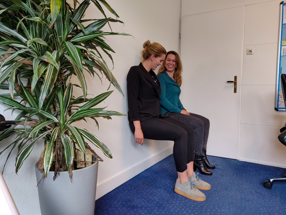 Foto van medewerkers die de oefening wall-sit uitvoeren op kantoor. Ze zitten met hun knieën in 90 graden gebogen tegen de muur aan.