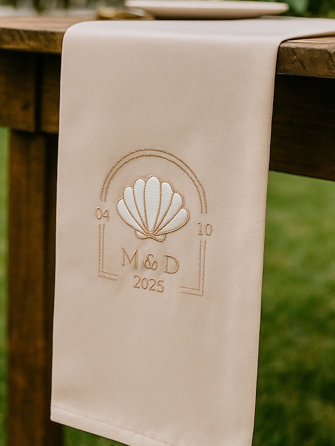 Embroidered wedding napkin featuring a shell motif, initials, and date, perfect for special occasion.