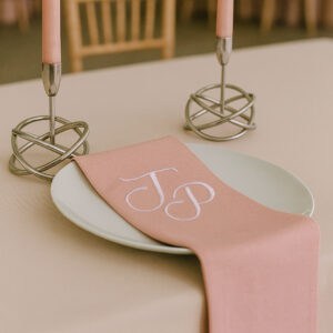 Embroidered pink napkin with white monogram initials "J & P" on a white plate, set on a dining table.