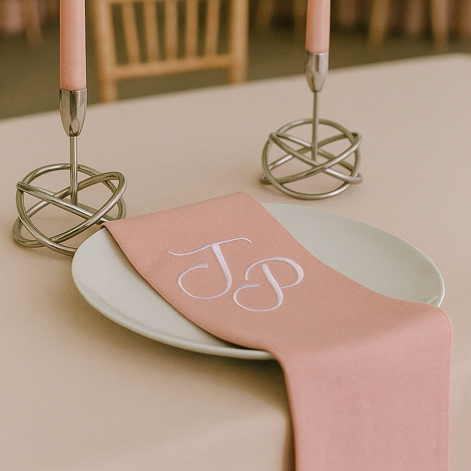 Embroidered pink napkin with white monogram initials "J & P" on a white plate, set on a dining table.