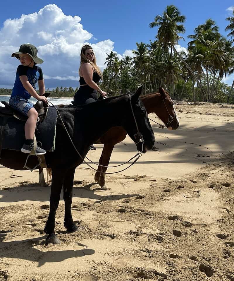Punta Cana Two women riding horses along the sandy Punta Cana shoreline with palm trees and blue sky.
