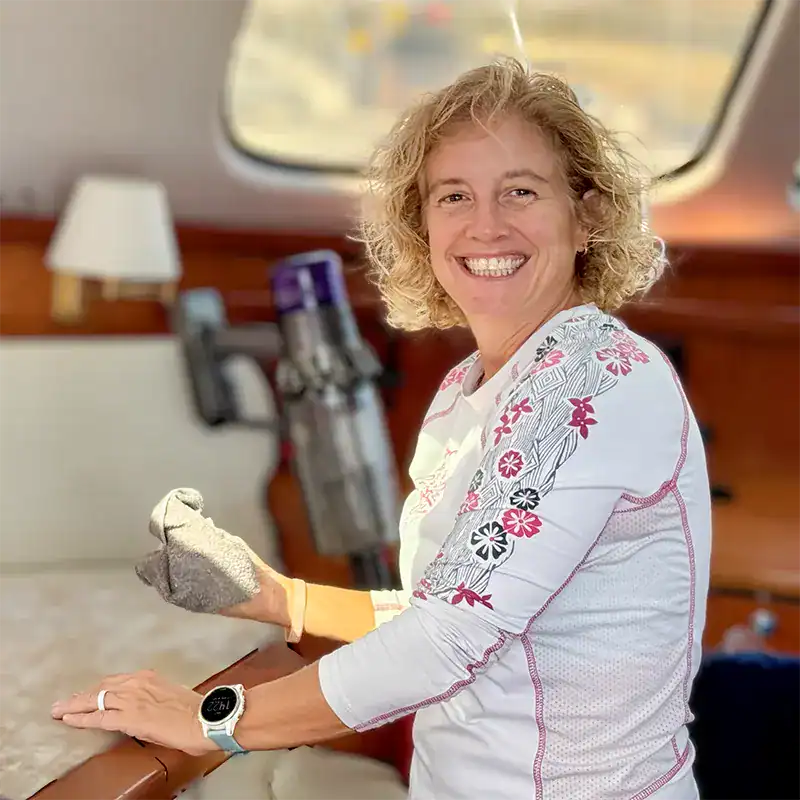 2. Woman smiling inside a sailboat, wearing gloves, with a cleaning cloth in hand, aboard an Antares Catamaran.