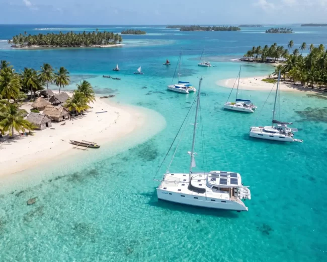 An elegant white catamaran floating in clear, calm Caribbean waters near a tropical island with palm trees and thatched-roof huts, perfect for serious cruisers seeking adventure and relaxation.