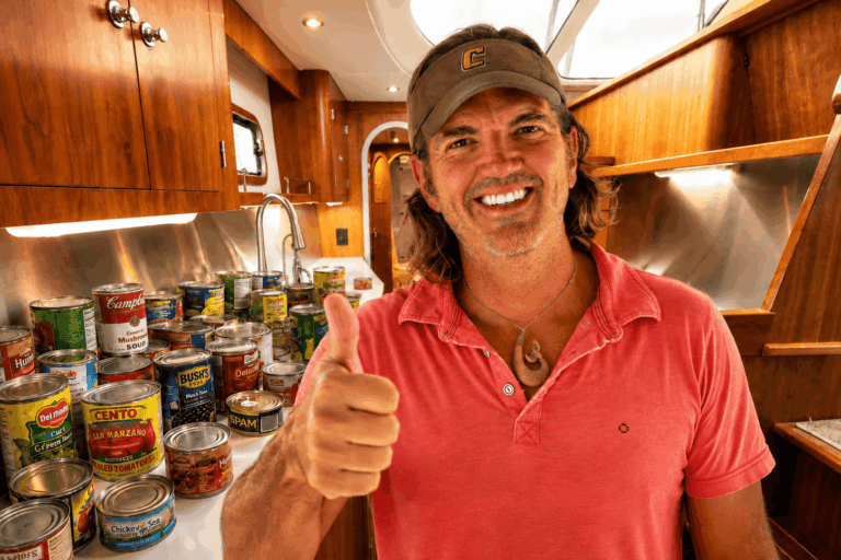 A man smiling and giving a thumbs-up inside a yacht kitchen with wooden cabinetry and canned food supplies.
