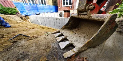 Close-up of an excavator bucket at a commercial construction site, with soil and debris around. A partially built retaining wall and building with blue tarp in the background hint at ongoing HVAC installations.