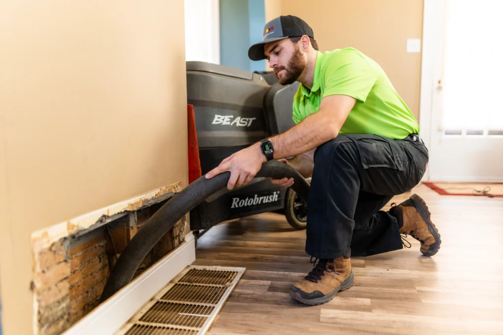 A HVAC technician in a green Anton's shirt and cap uses a large vacuum hose to perform air duct cleaning through an opening in the wall of a St. Louis residential home during summer.